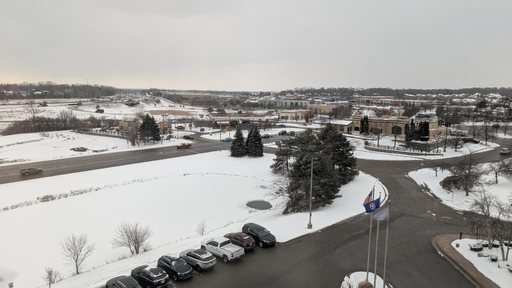 The view from the hotel window at ConFusion 2025. A parking lot, a frozen pond, and several roads are visible, as well as various evergreen and deciduous trees.